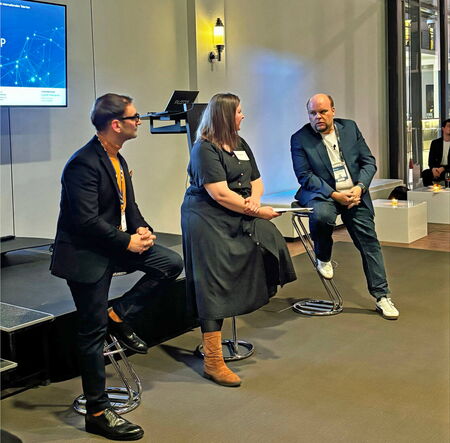 Three people are seated on bar stools on a stage during a panel discussion. Two men and one woman are engaged in conversation. Behind them, a screen displays a presentation, and a lectern stands nearby. The room has a modern setting with dim lighting.