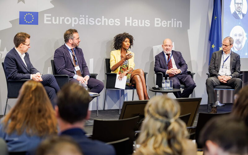 Vier Diskussionsteilnehmer und die Moderatorin sitzen in einem Halbkreis auf der Bühne, und die Moderatorin in der Mitte hält die Einführungsrede zur Veranstaltung.