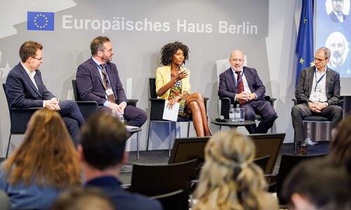 Four panelists and the moderator sit on stage in a semicircle and the moderator at the center is doing the intro to the event.